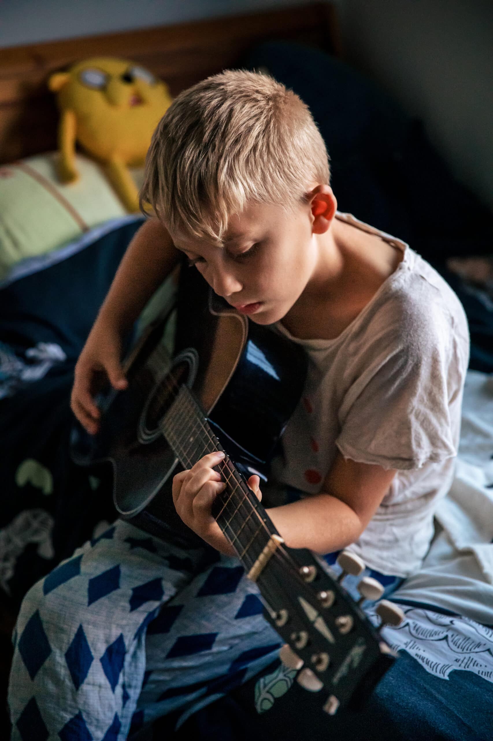 Blond child practicing acoustic guitar on a cozy bedroom bed in natural light.