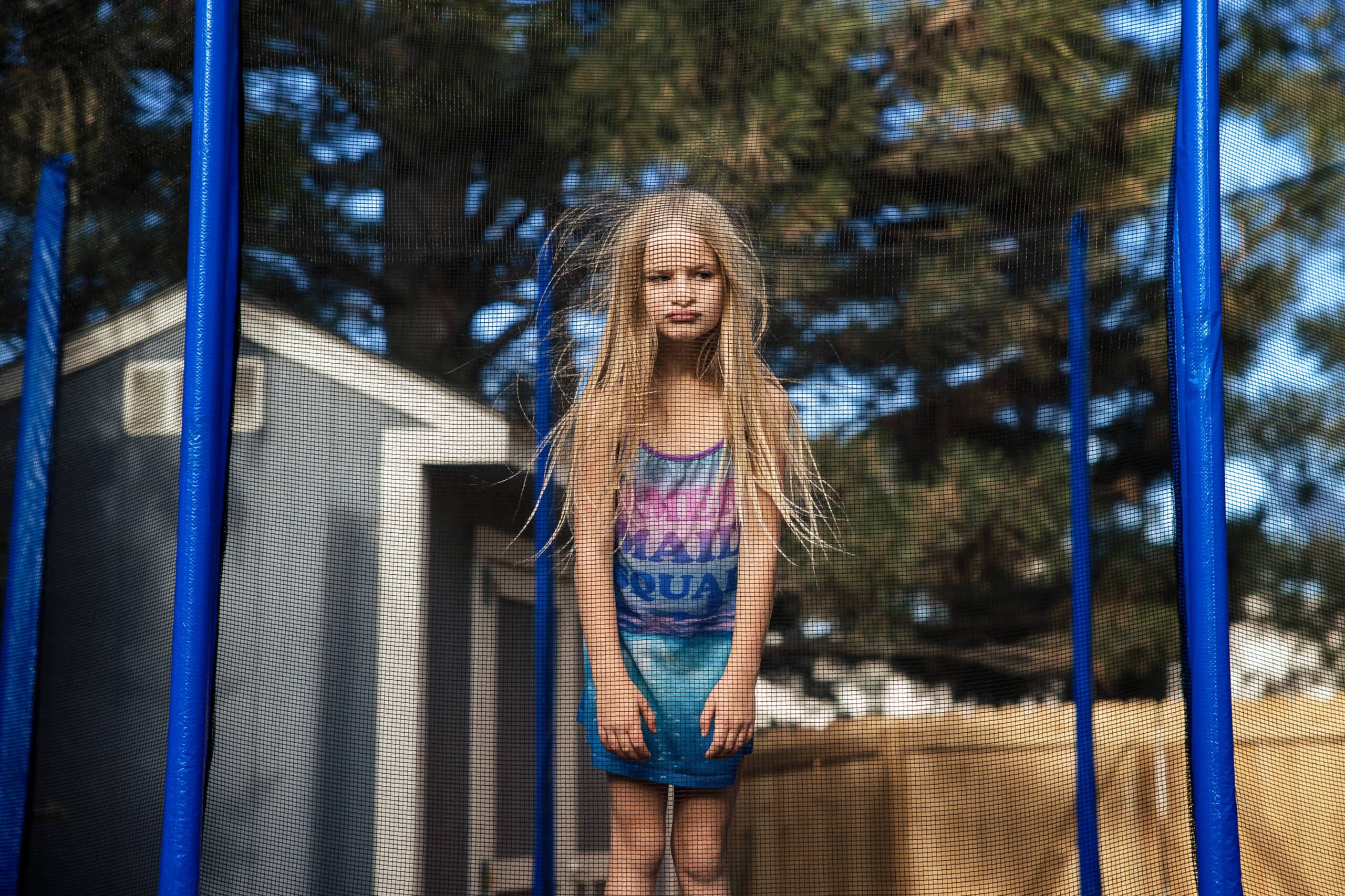 Blonde girl framed by trampoline poles in a sunny backyard portrait.