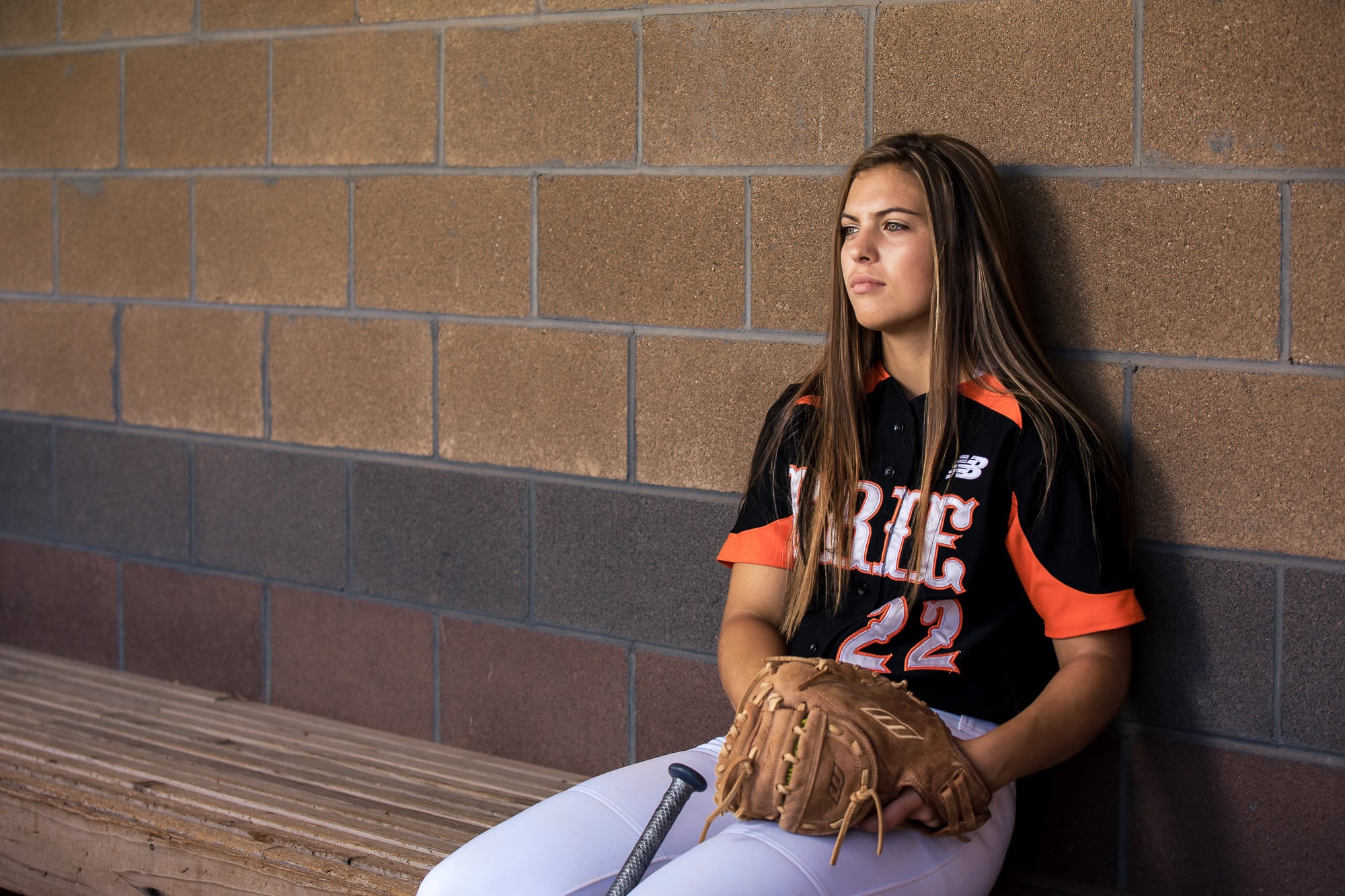 A high school senior sitting in the softball dugout with a baseball bat and glove.