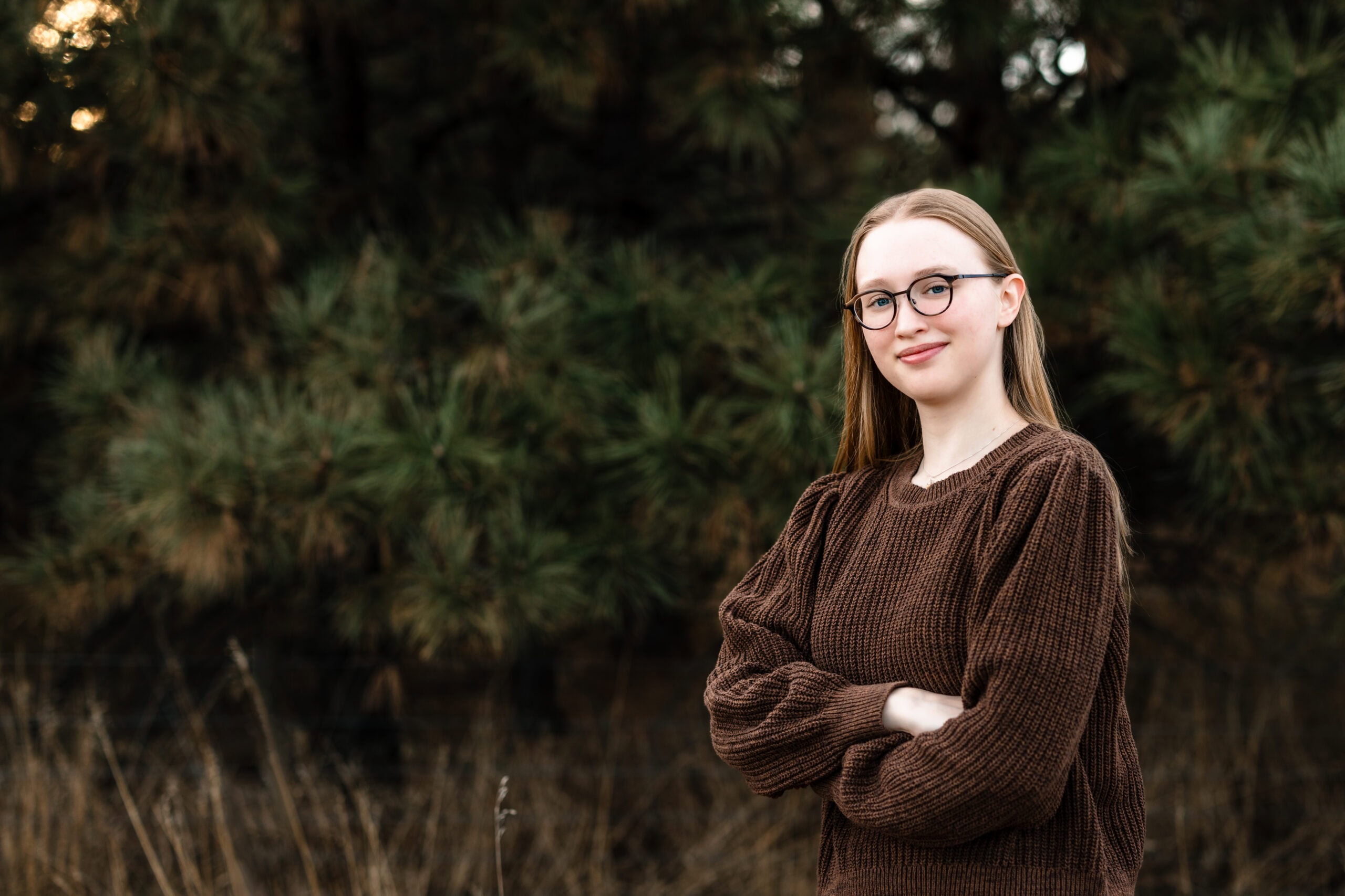 High school senior pictures session of a girl wearing a brown sweater on a background of dark fir trees.