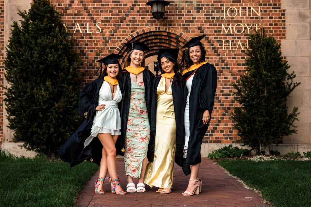 Colorado Senior Pictures of four friends in graduation gowns smiling under a brick arch