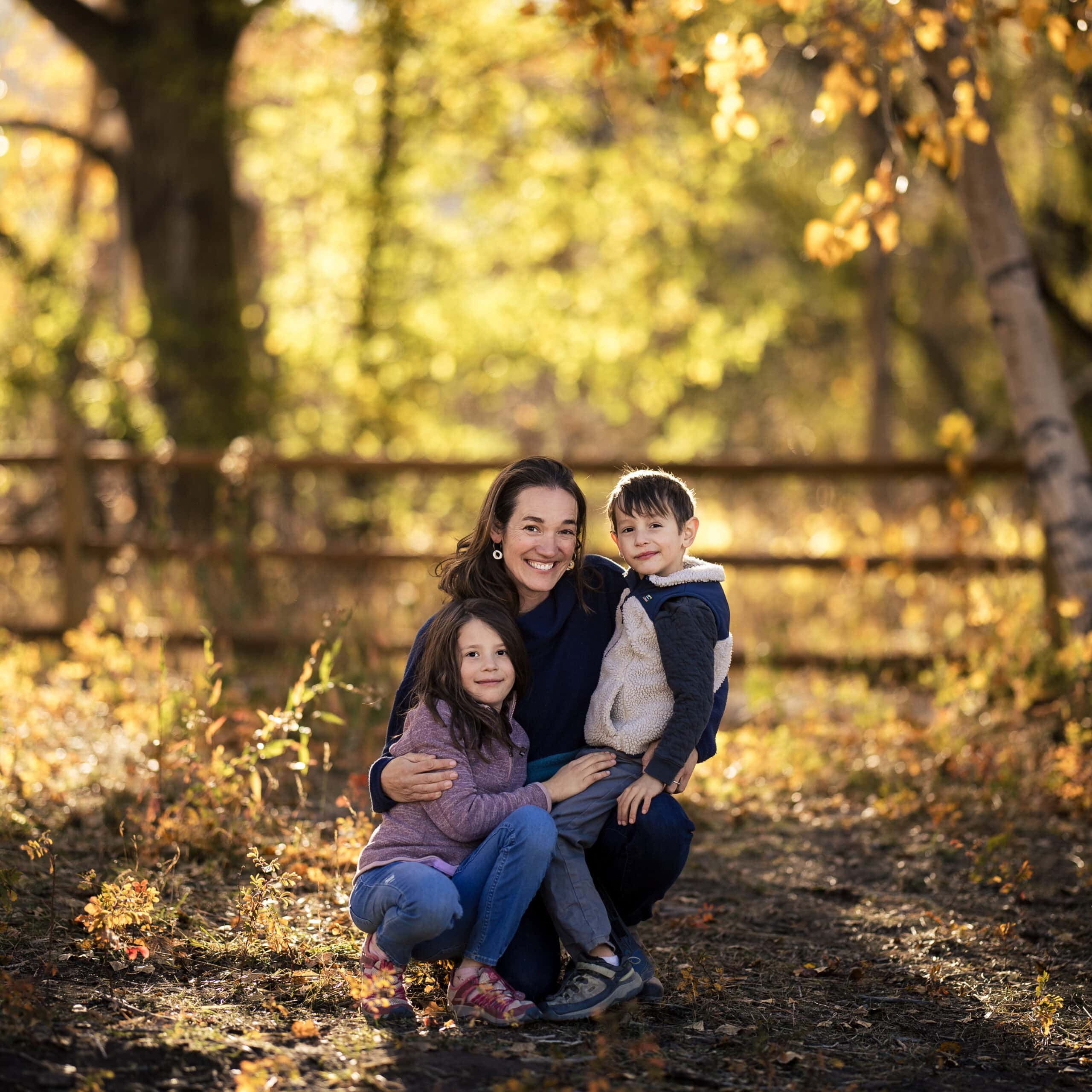 Mother hugging two smiling children on an autumn path.