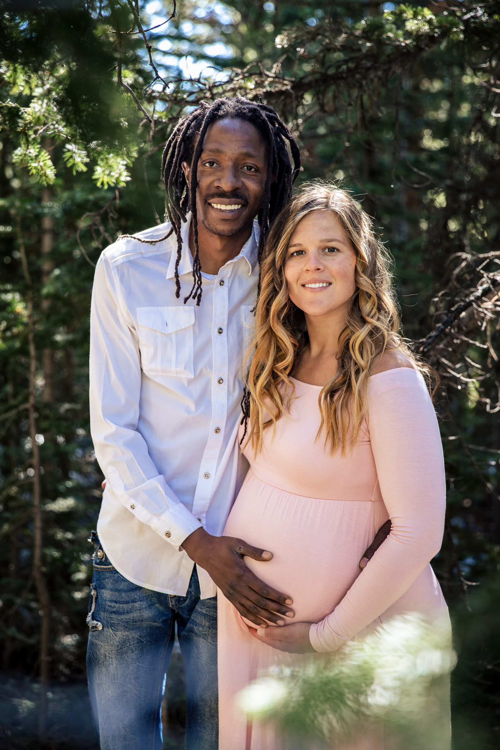Boulder maternity photographer captures smiling couple cradling baby bump in sunlit woods.