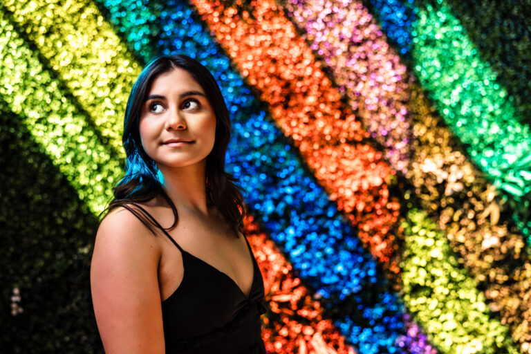 Graduate Mini Sessions portrait of senior in black top before glittering rainbow wall.